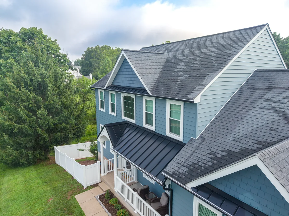 Closer aerial view across a blue home’s roof, porch, and front yard.