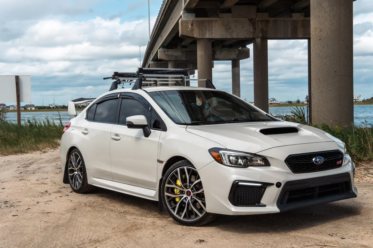 A white performance sedan photographed beneath a concrete bridge beside the water.