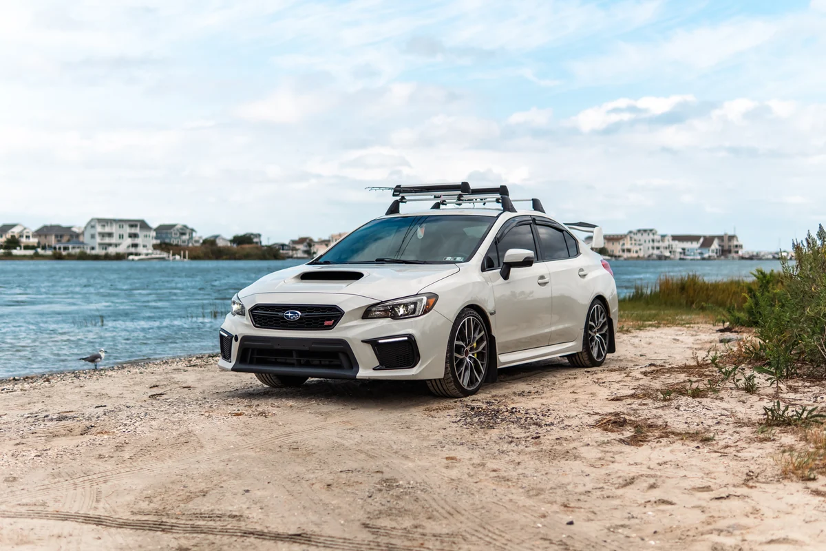 A white performance sedan parked beside the shoreline on a sandy path.
