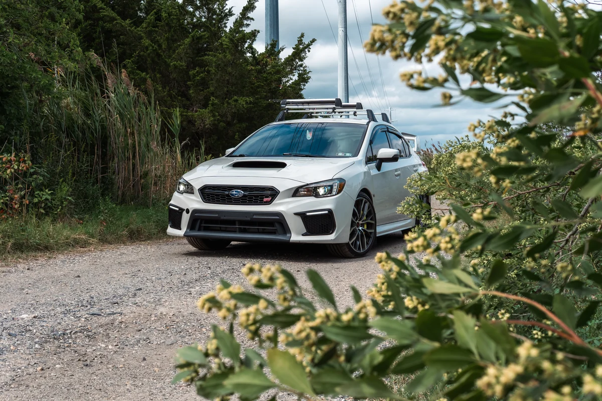A white performance sedan framed by coastal greenery along a sandy path.