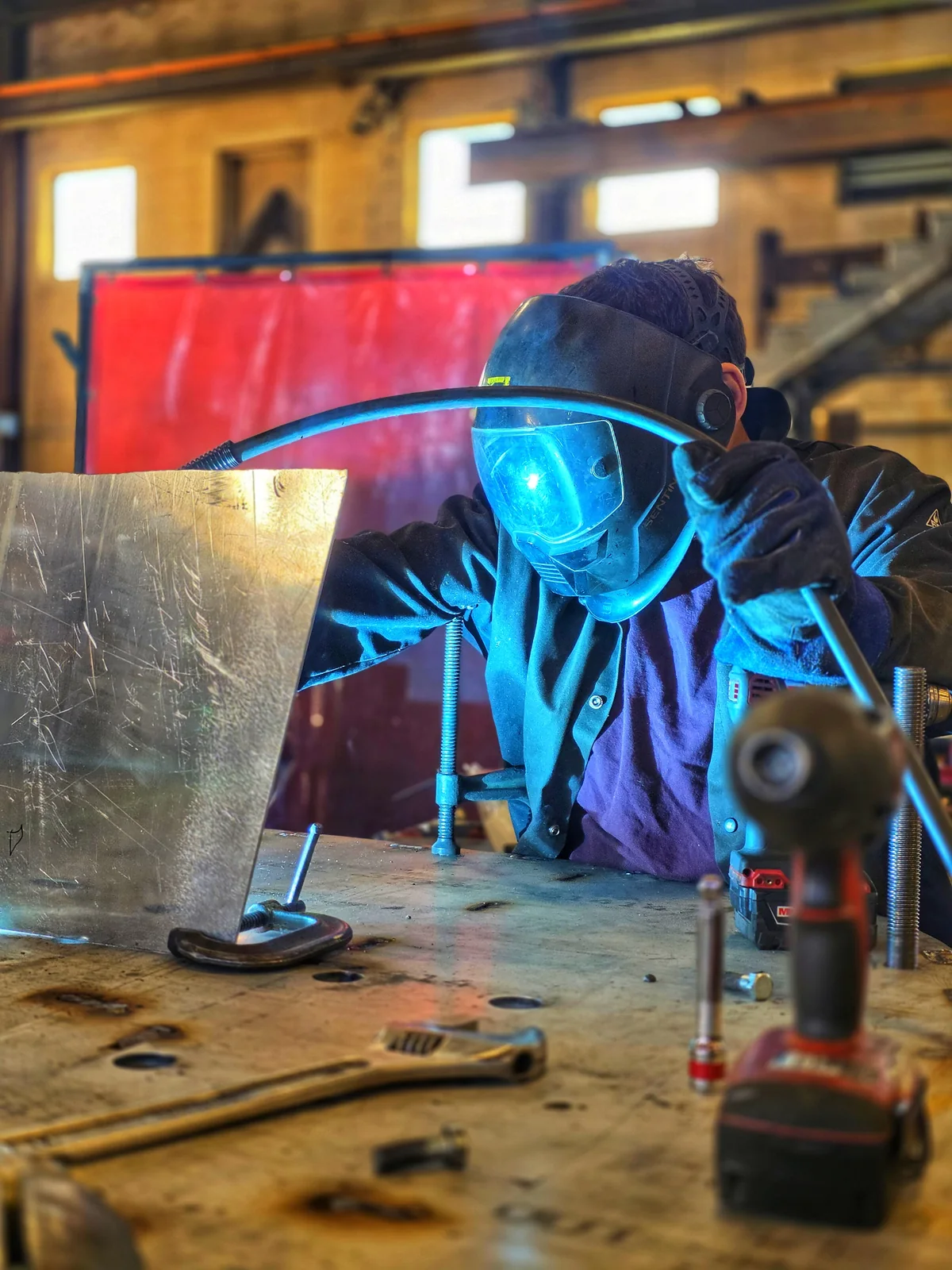A welder works behind a protective shield at a fabrication table, lit by a blue welding arc.
