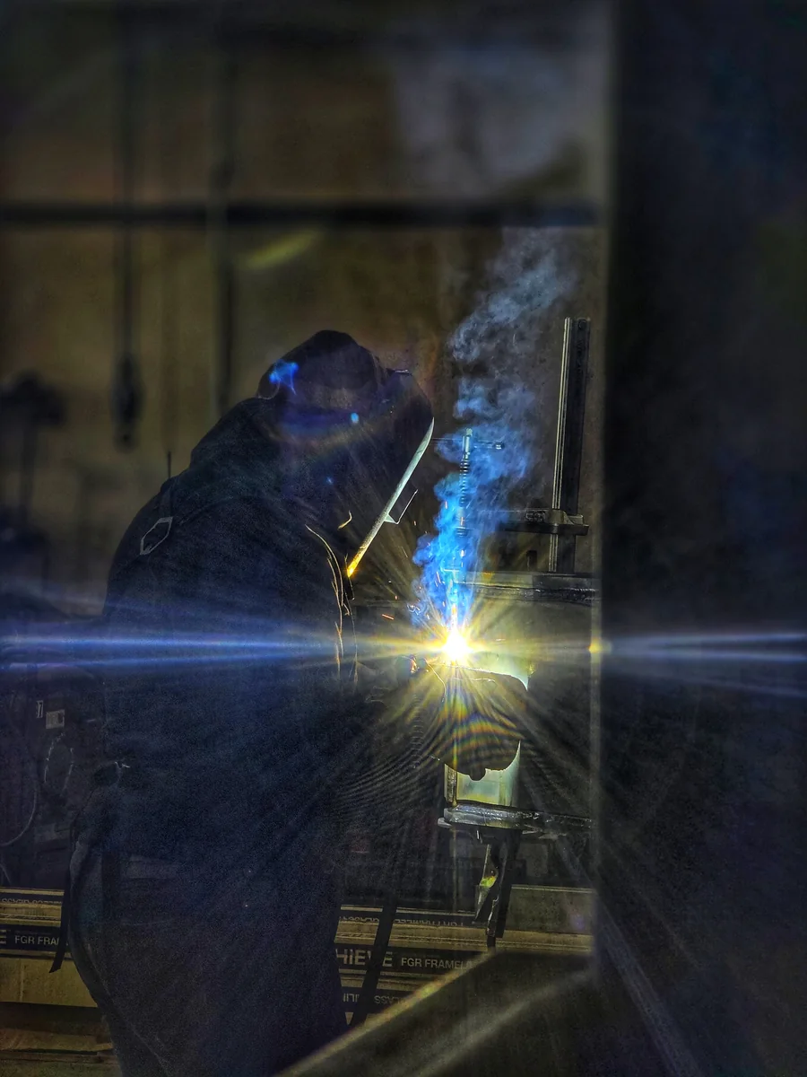 A welder works on a steel assembly inside a workshop, with a blue arc illuminating the scene.