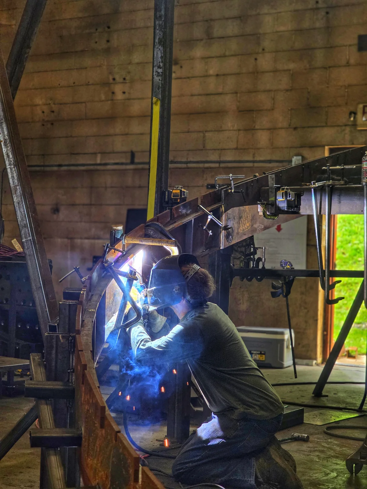 A welder joins a large steel frame inside a workshop.