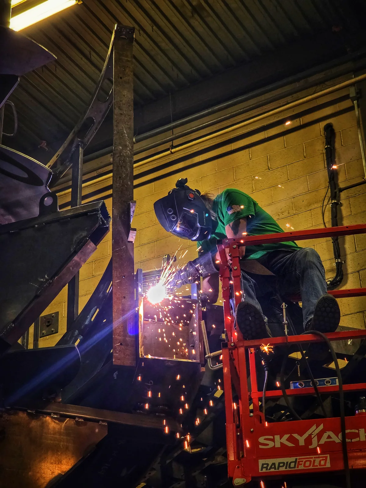 A welder works from an elevated platform beside a steel structure as sparks fall.