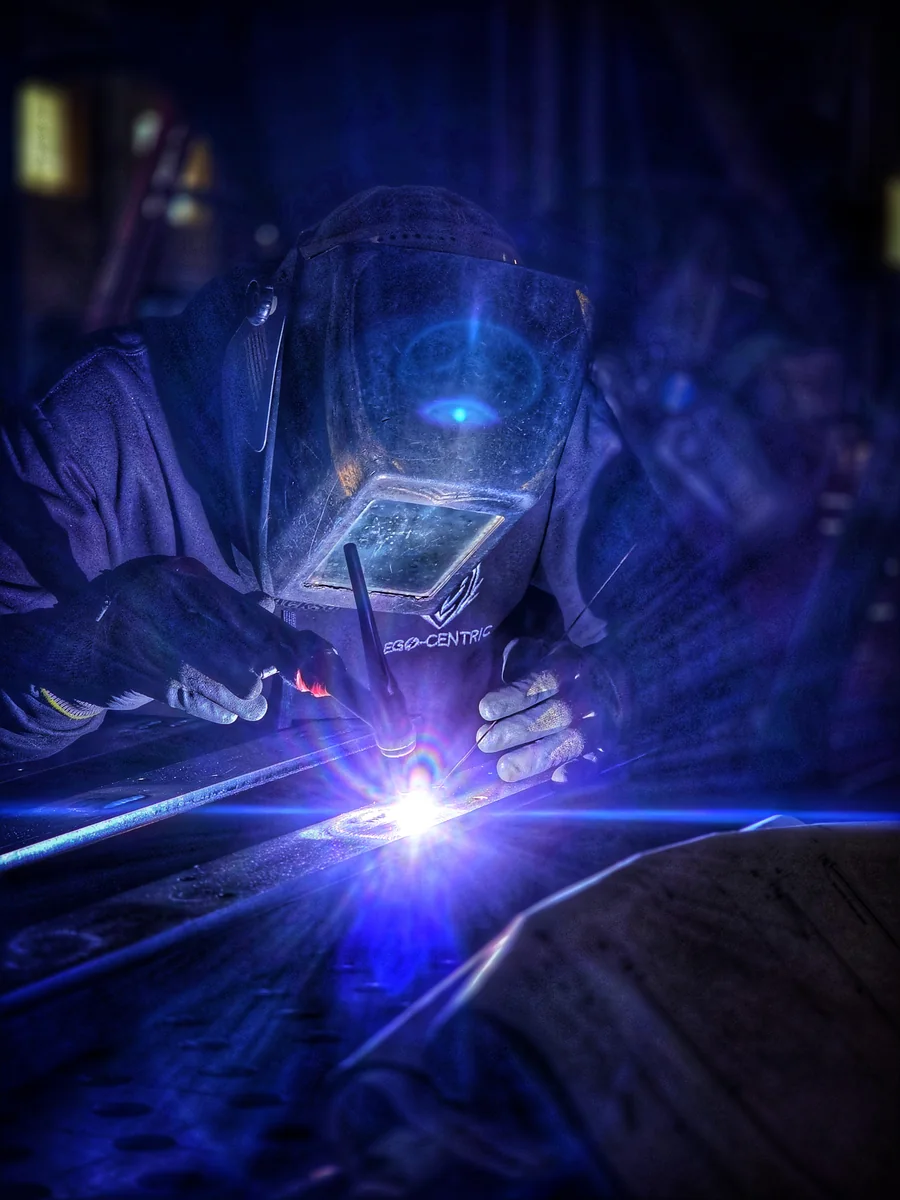 A welder in a protective helmet works close to a blue arc on a metal piece.