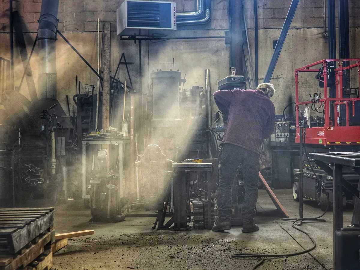 A worker shapes metal in a fabrication shop filled with tools, lifts, and industrial equipment.