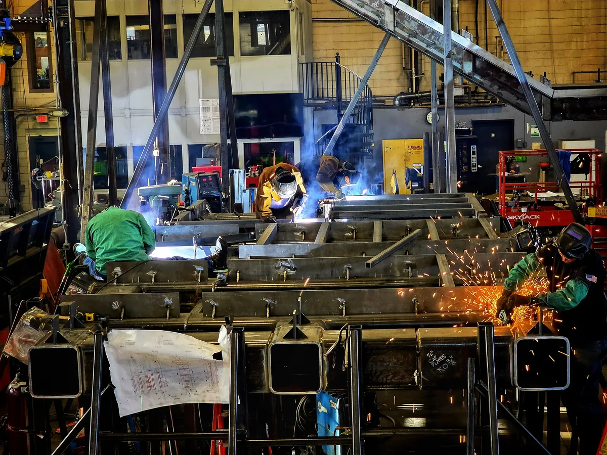 Several welders fabricate a large steel structure inside an industrial workshop.